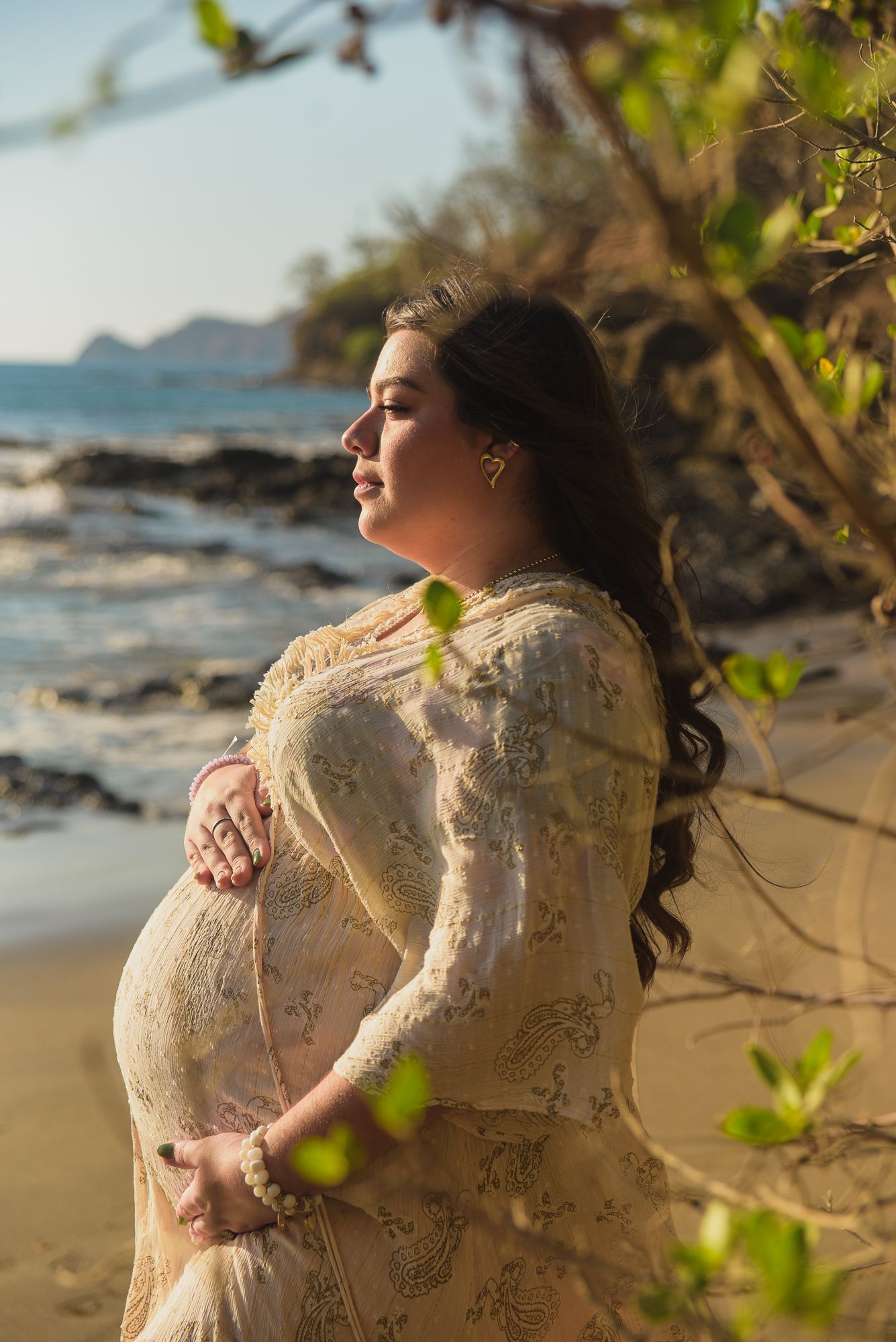 Pregnant woman in lace dress cradling baby bump on rocky beach at golden hour at four season, Papagayo, Costa Rica