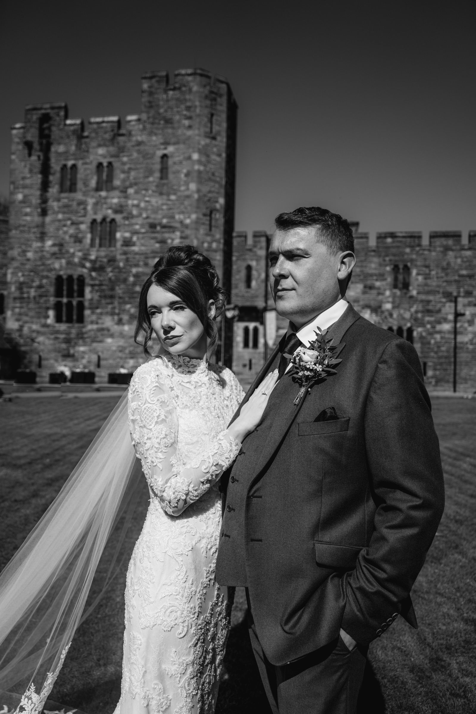 Black and white wedding portrait of a bride in a lace gown and veil beside her groom in a suit, with a historic stone castle behind them.
