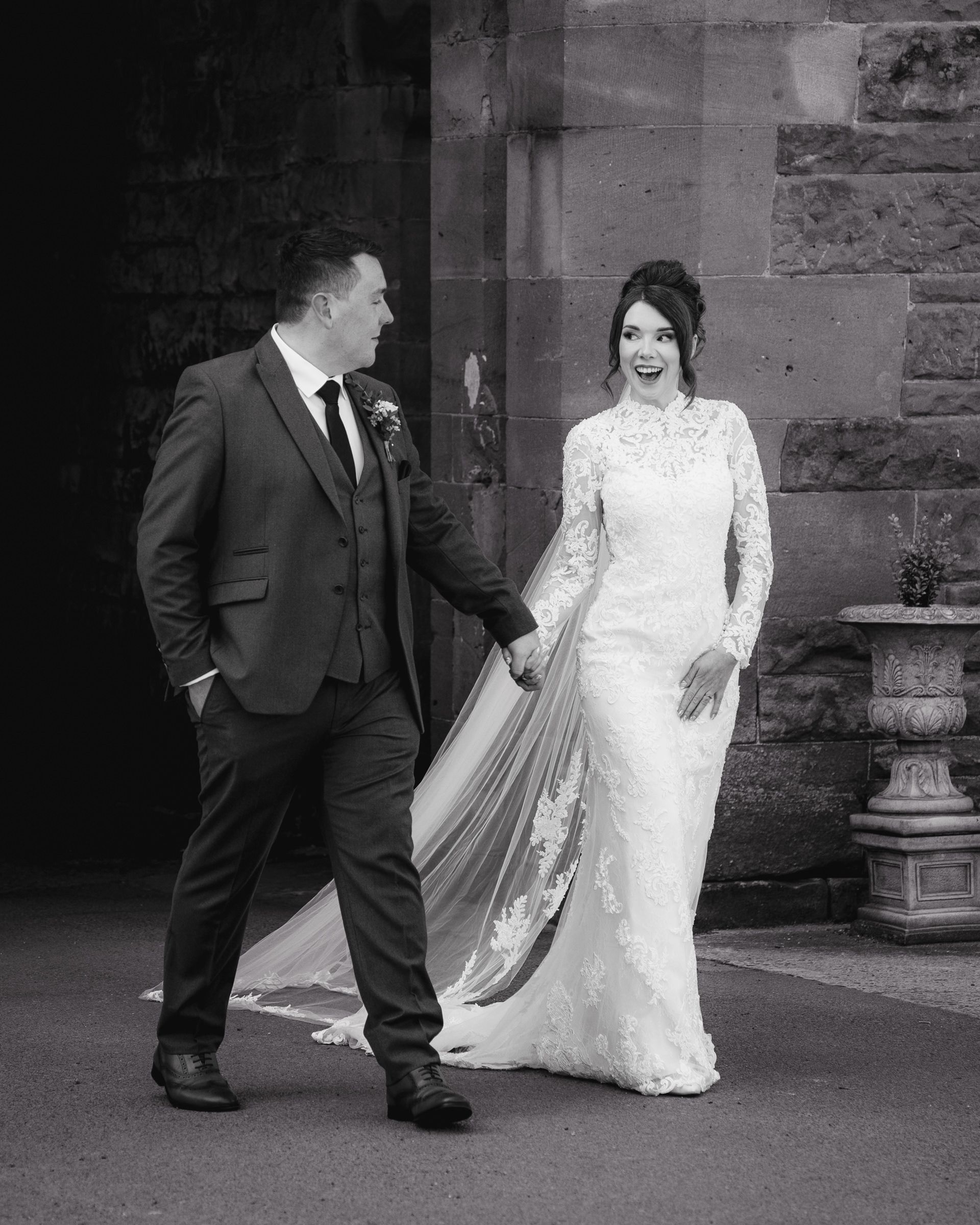 Black and white photo of a smiling bride in a lace gown with a long veil walking hand in hand with a groom in a suit by a stone wall.