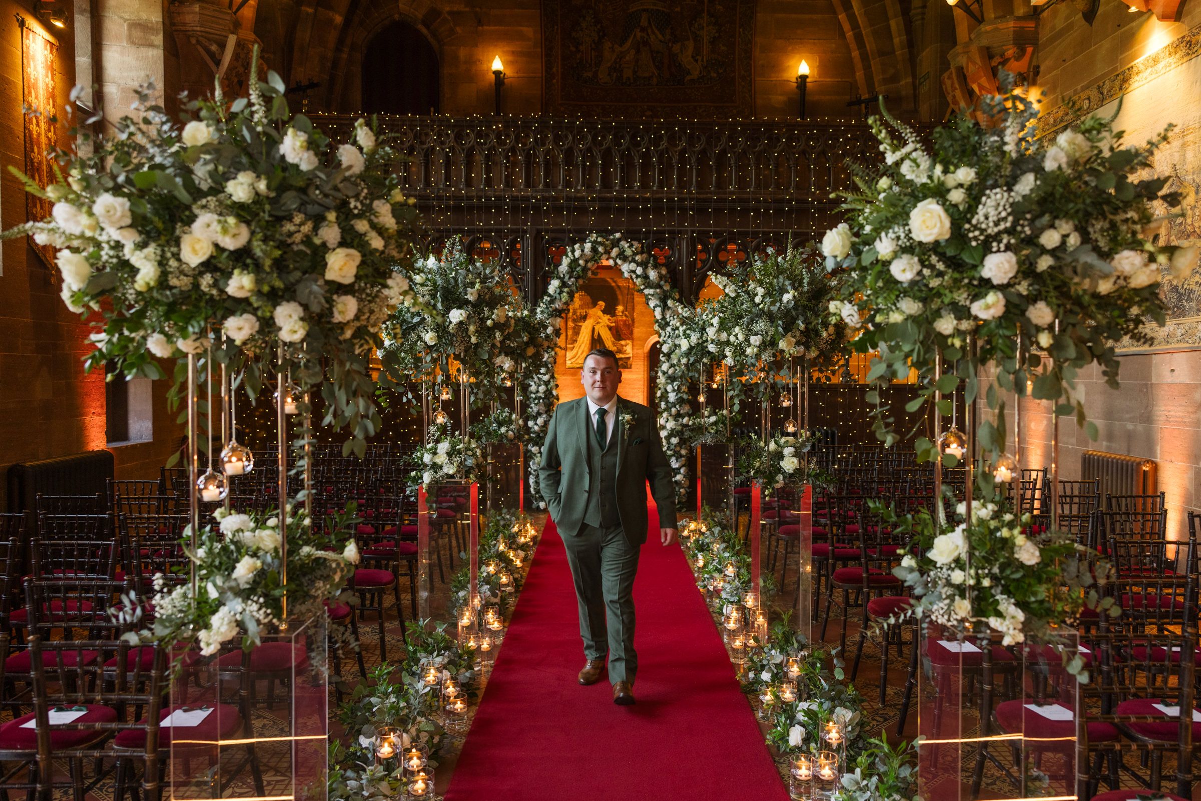 Groom in a dark suit walks down a red carpet aisle lined with candles and white floral arrangements in an ornate church wedding venue.