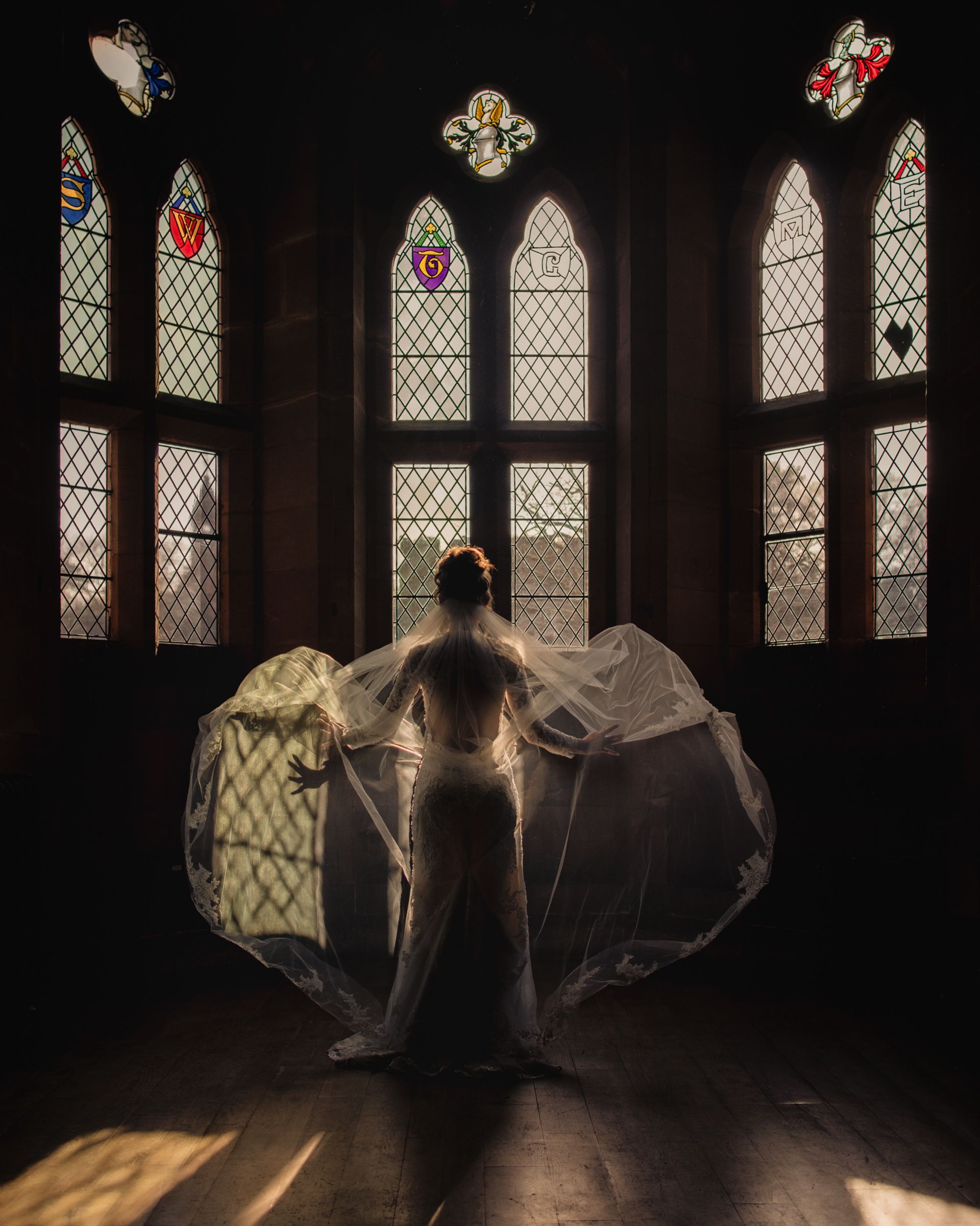 Bride in a flowing white veil and gown stands silhouetted before Gothic stained glass windows as sunlight streams across a wooden floor.