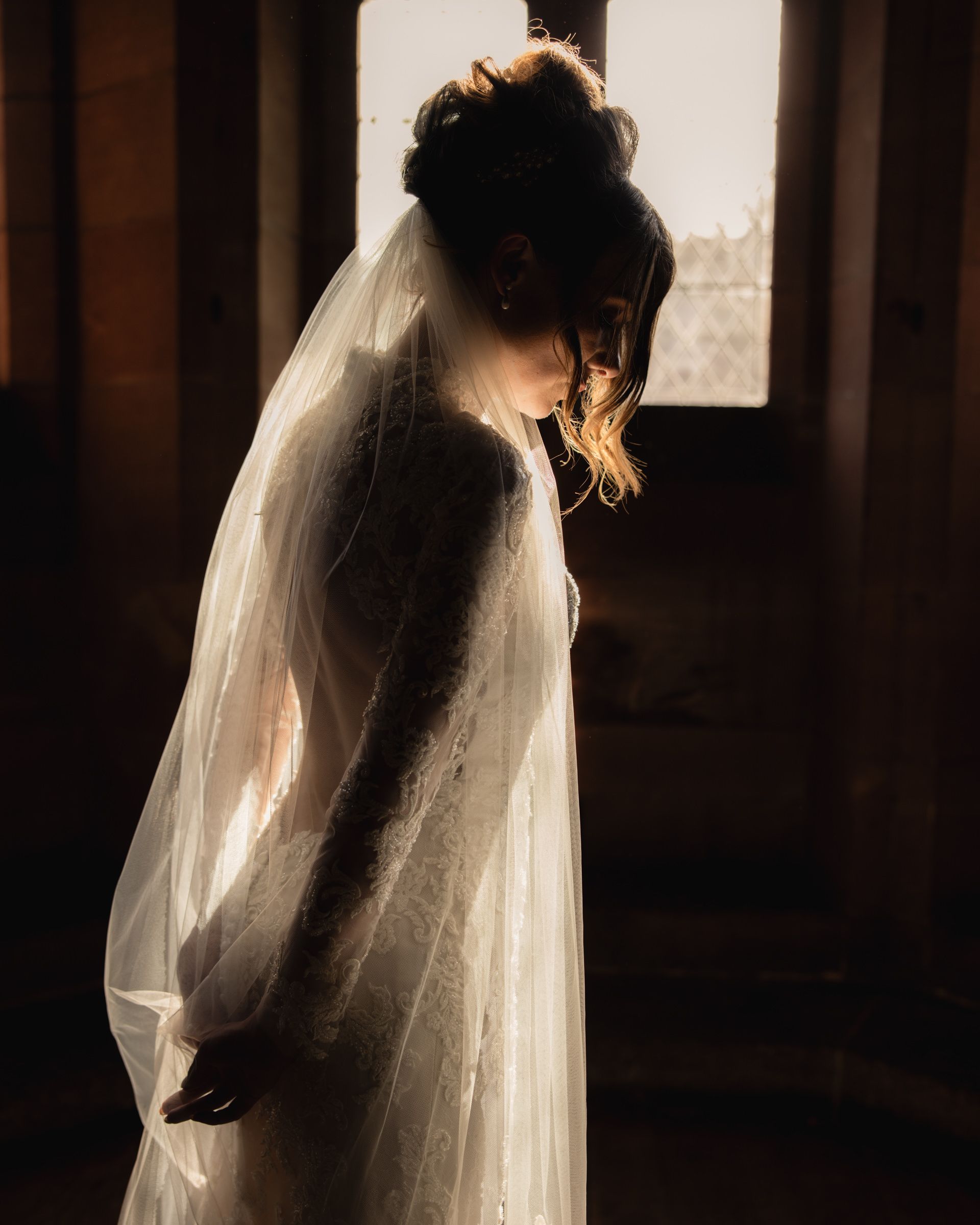 Backlit bride in a long-sleeved lace wedding gown and sheer veil, head bowed, standing by a sunlit window in a softly shadowed room.