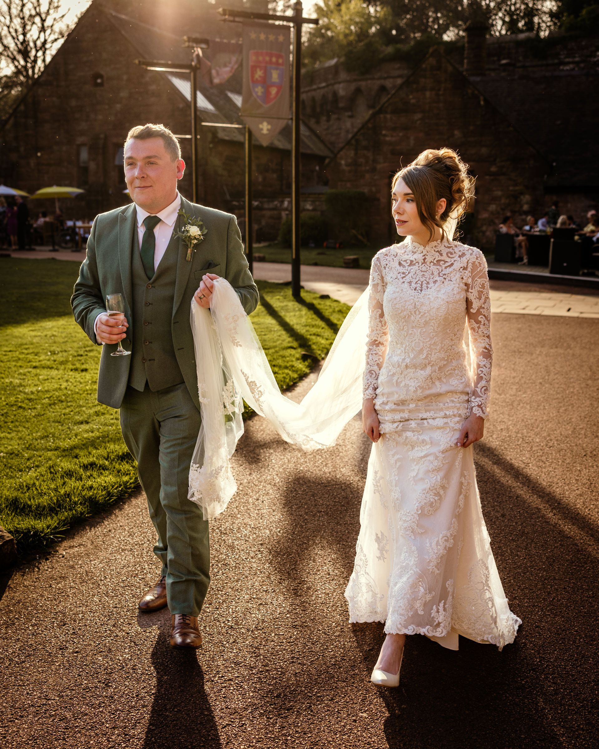 Groom in a green three-piece suit holds a drink and the bride's veil as they stroll a sunlit path in a lace gown at a wedding.