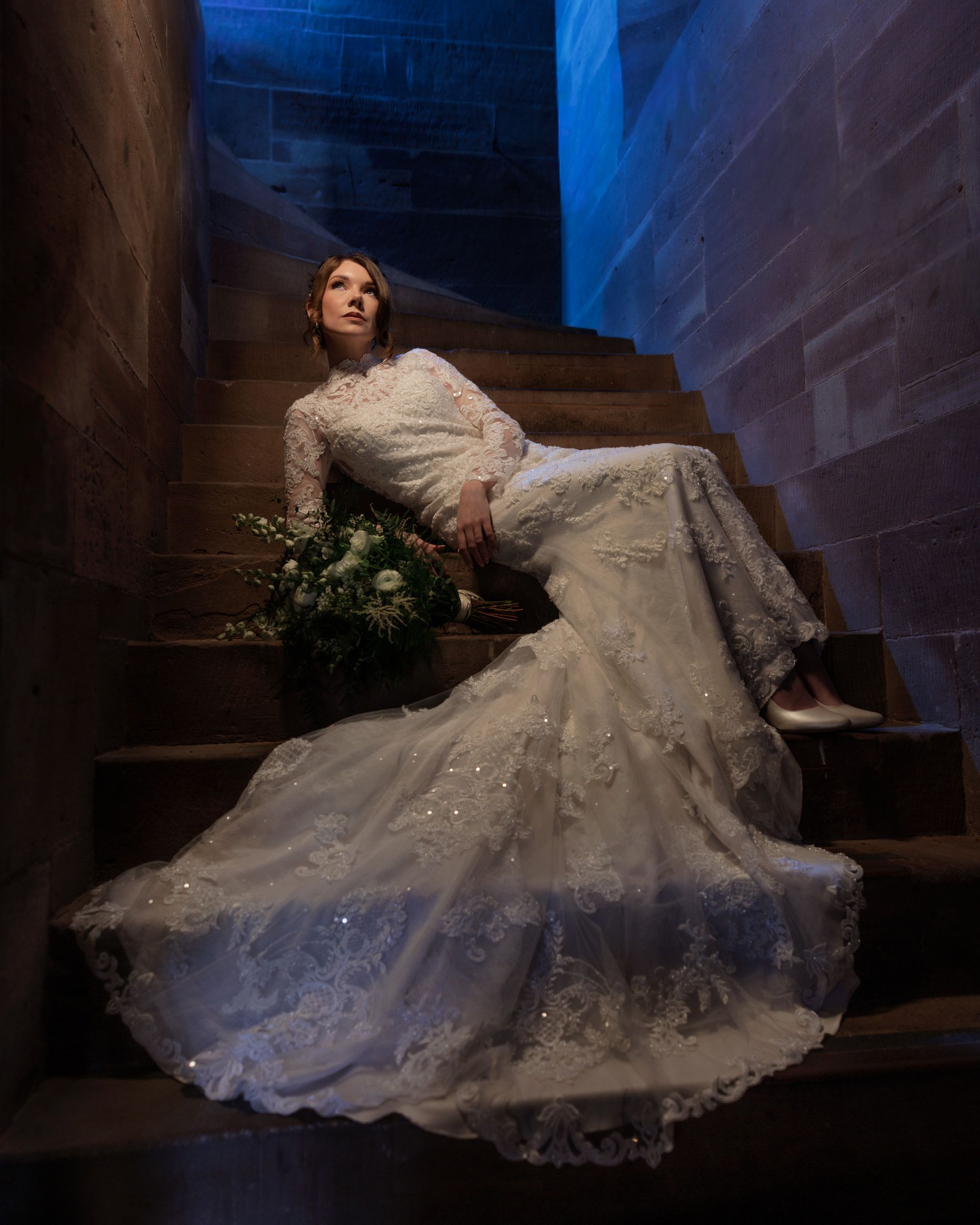 Bride in an elegant lace wedding gown reclining on dimly lit stone stairs while holding a white floral bouquet.