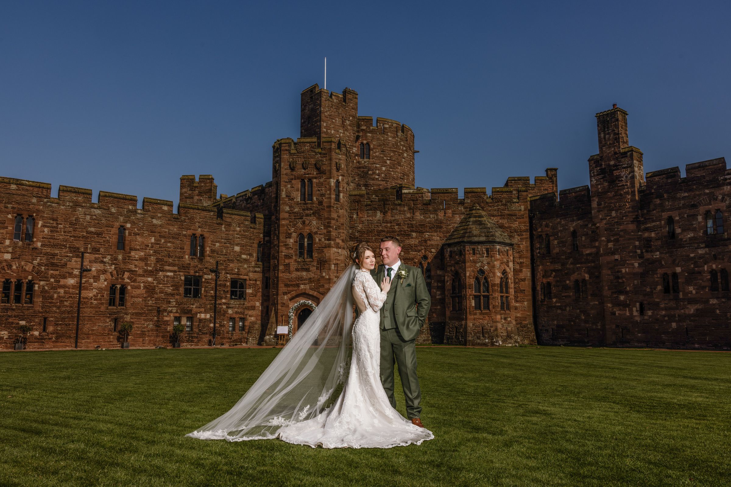 Bride in a flowing white gown and veil poses with groom in a green suit on a lawn in front of a historic red sandstone castle at sunset.