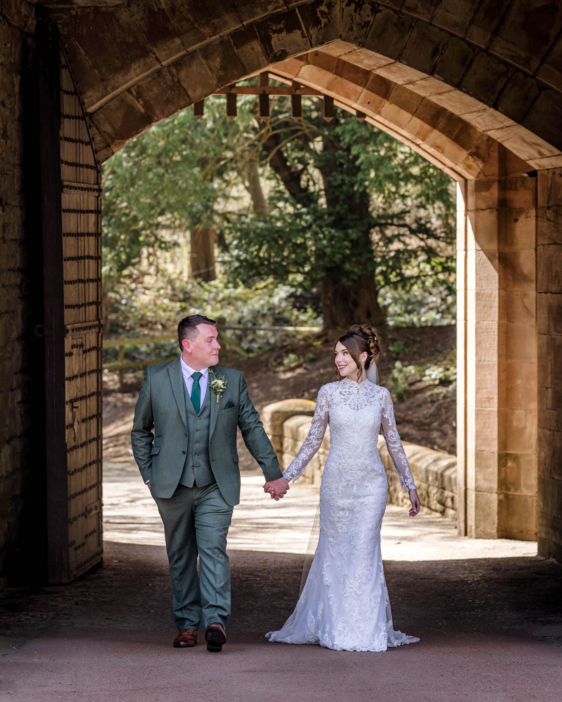 Bride in a lace wedding gown and groom in a green three-piece suit hold hands walking through a stone archway toward sunlit woodland.