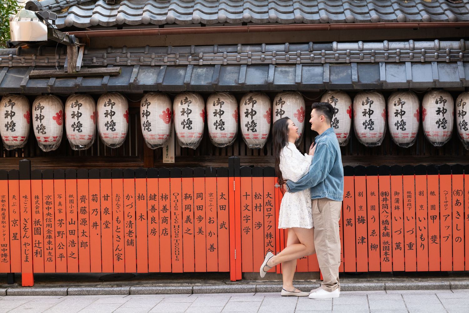 Couple embracing in front of an orange wooden fence with rows of white Japanese paper lanterns hanging above a traditional tiled roof.