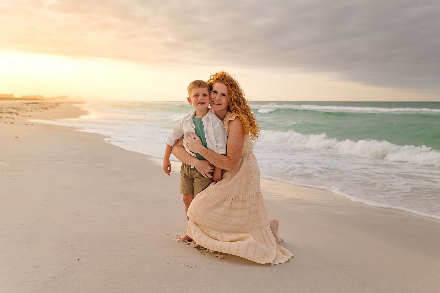 Woman in beige dress hugging young boy on sandy beach at sunset with ocean waves in background.