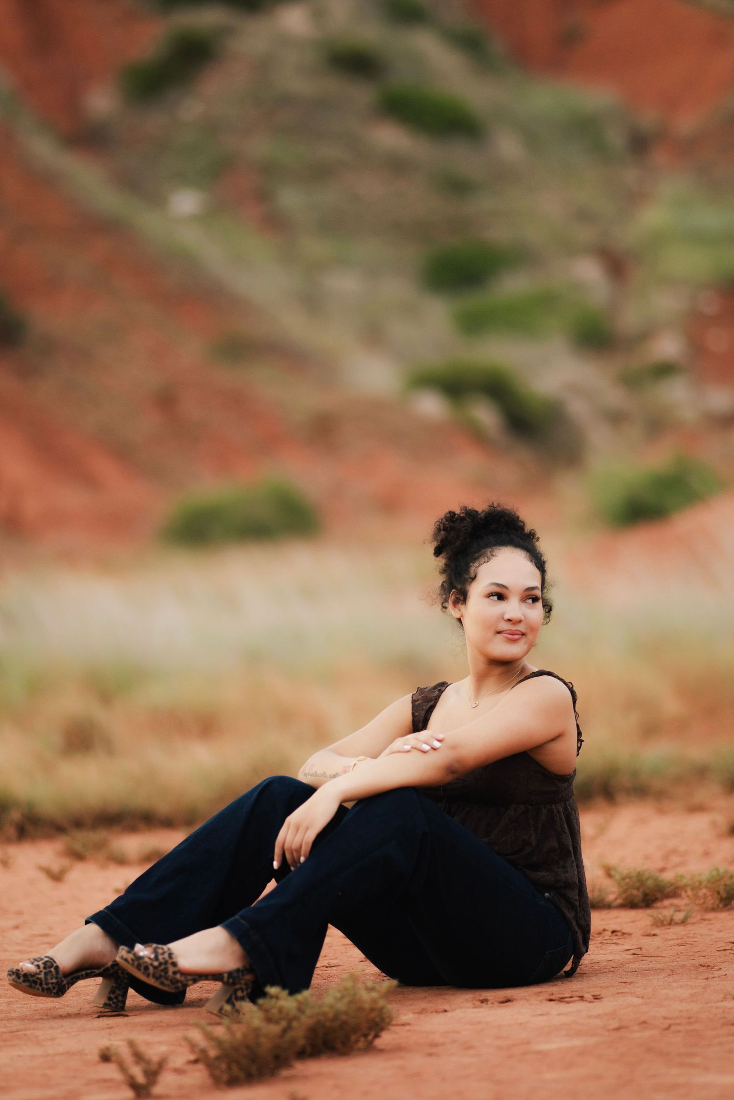 A young woman with curly hair sits smiling on red desert sand, wearing dark jeans and a sleeveless top.