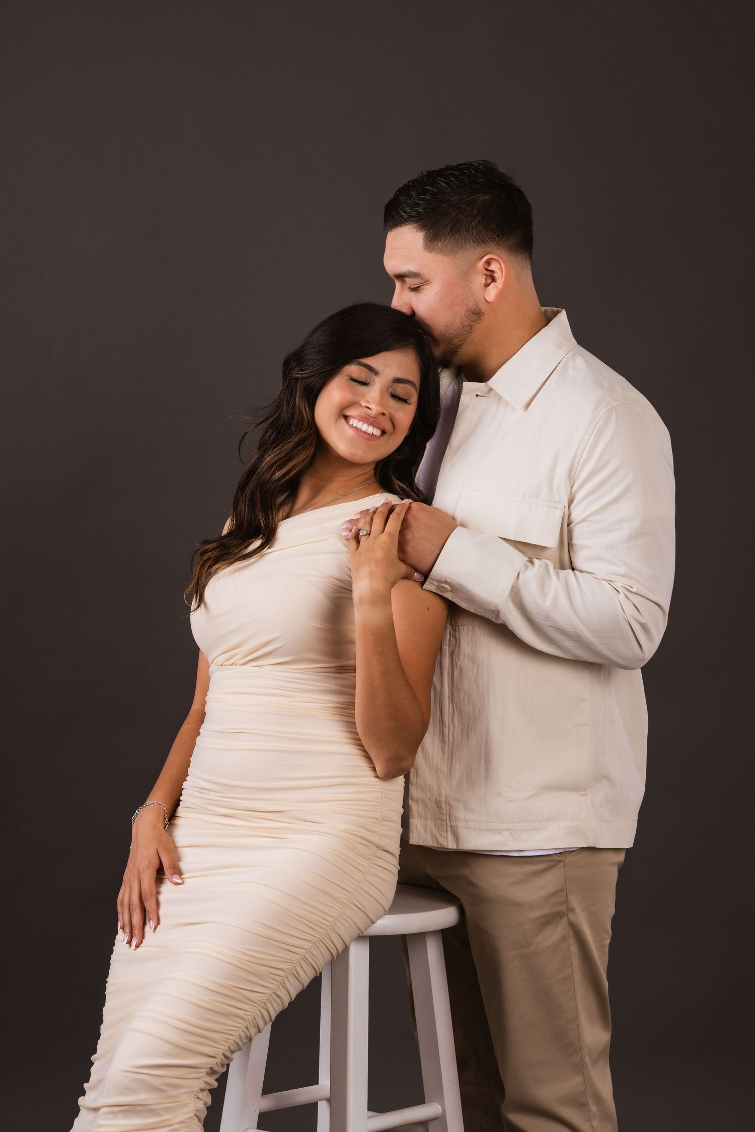 A couple poses intimately in a studio, woman in cream ruched dress leaning on stool, man in beige shirt embracing her.