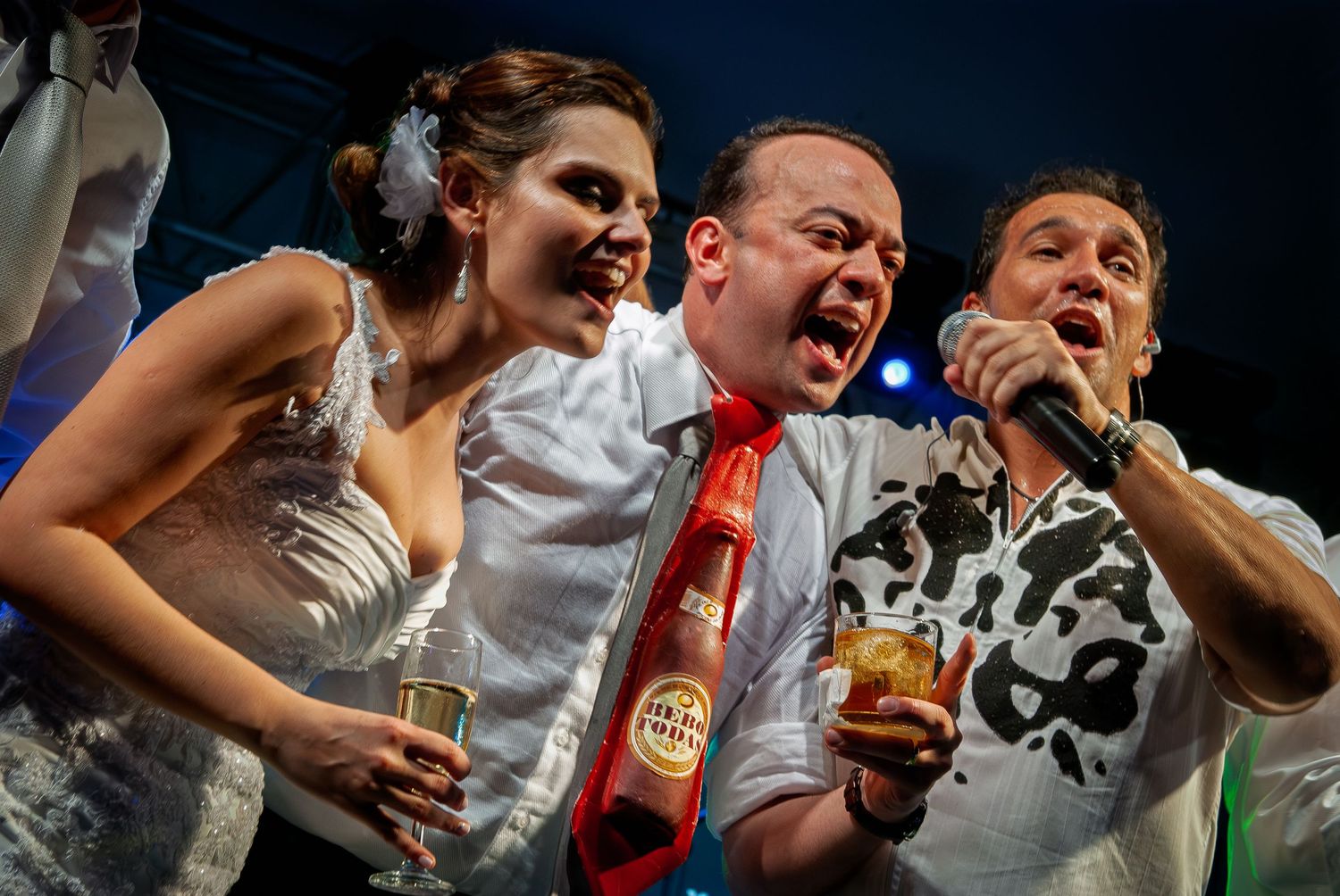 Bride and groom singing joyfully with a performer at a lively colorful wedding reception celebration.