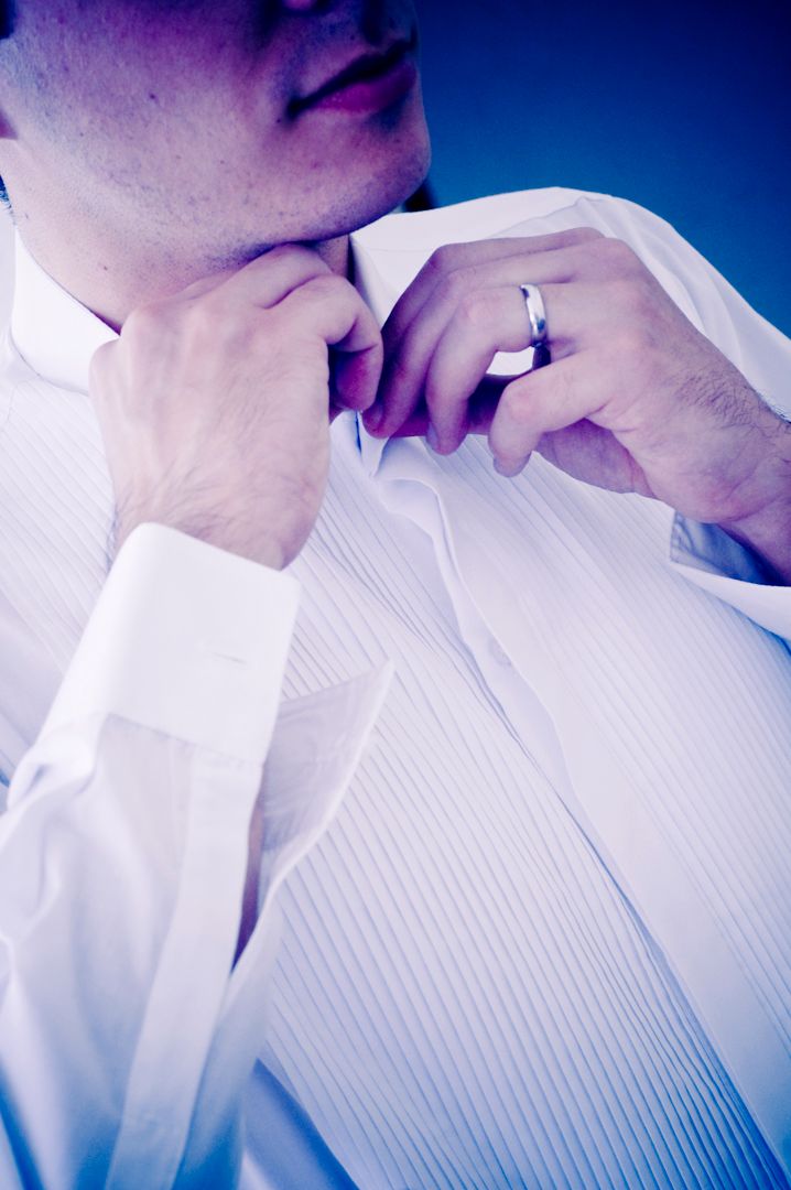 Man in white tuxedo shirt adjusting collar, wearing a silver ring, against blue background.