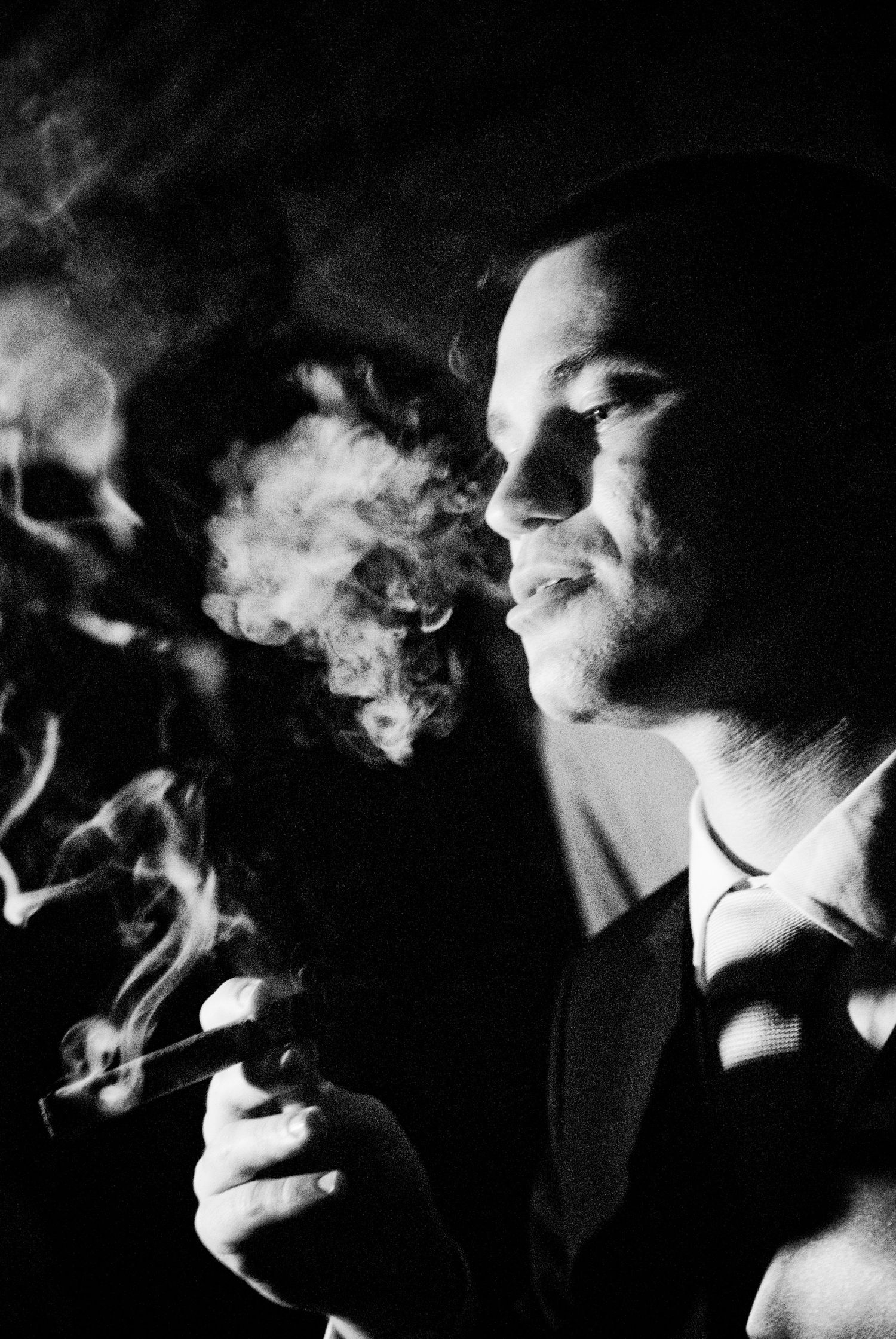 Dramatic black and white portrait of a groom smoking a cigar with swirling smoke in dark lighting.