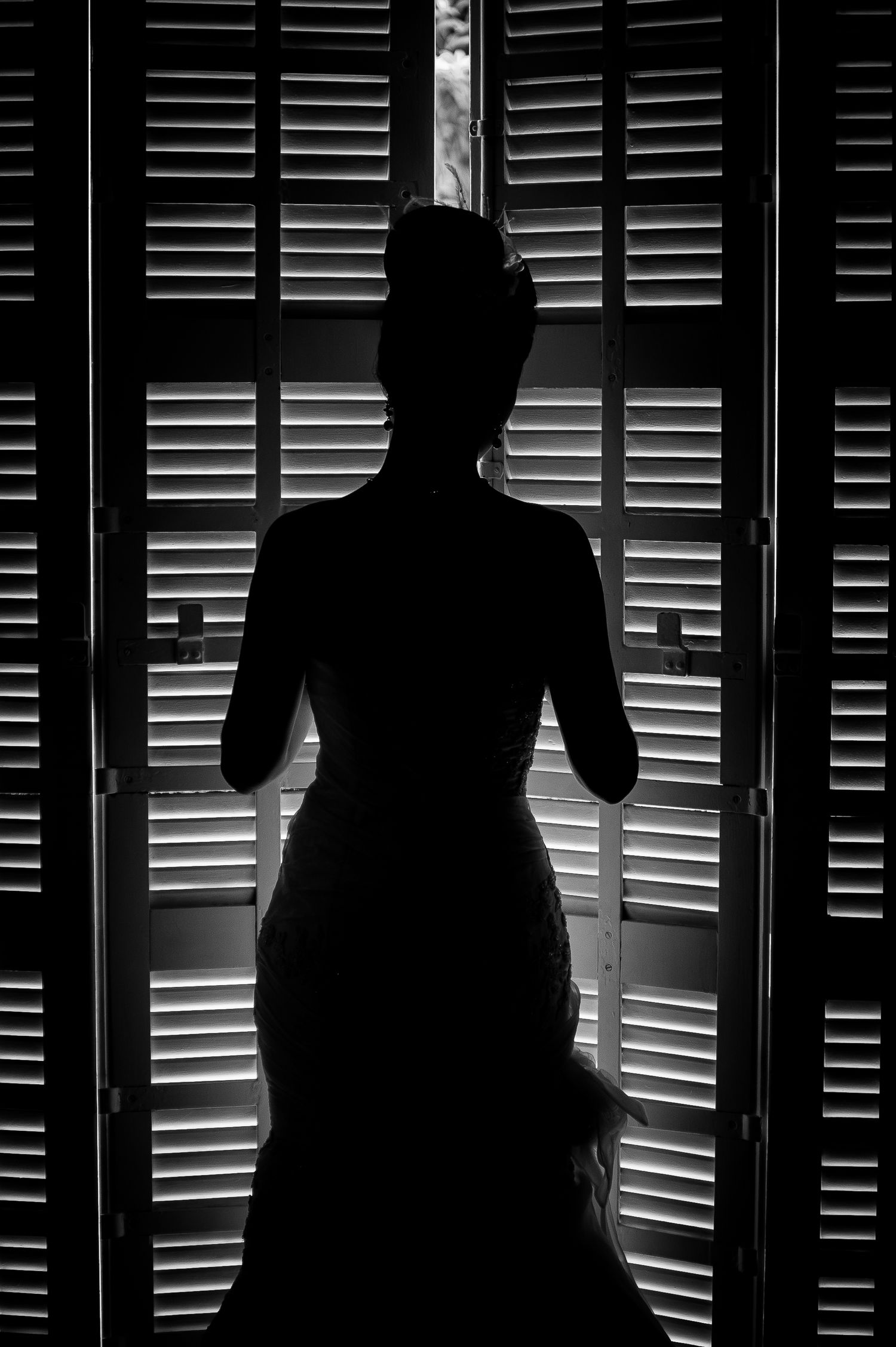 Silhouette of a bride in wedding gown standing before louvered shuttered doors in dramatic lighting.