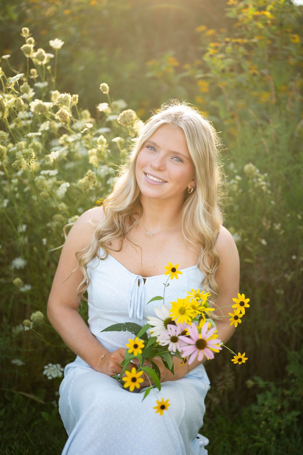 Smiling blonde woman in a light blue dress holding colorful wildflowers while sitting in a sunlit meadow.
