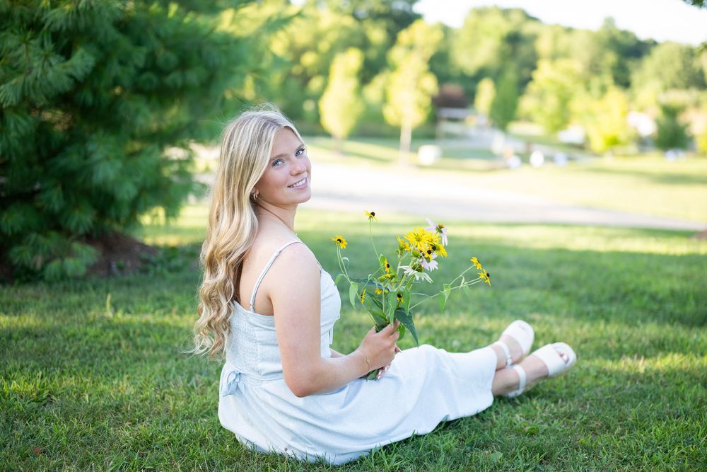 Smiling blonde woman in white dress sitting on grass holding yellow wildflowers in a sunny park setting.