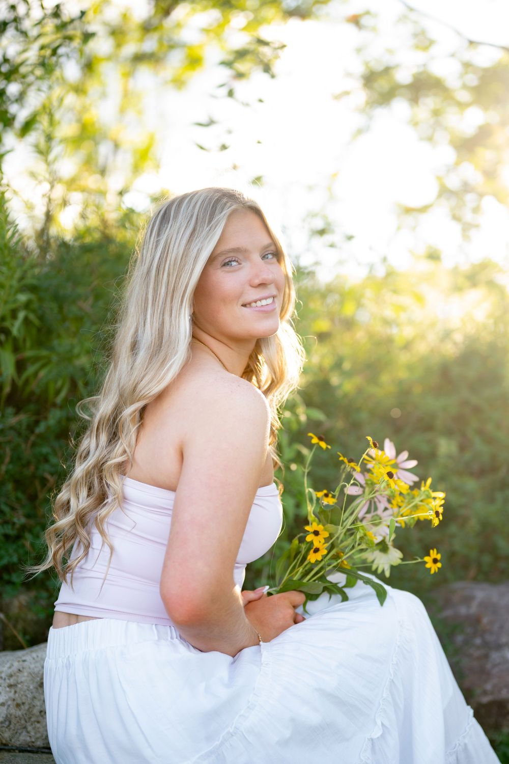 Smiling blonde woman in white strapless dress holding wildflowers outdoors in golden sunlight.