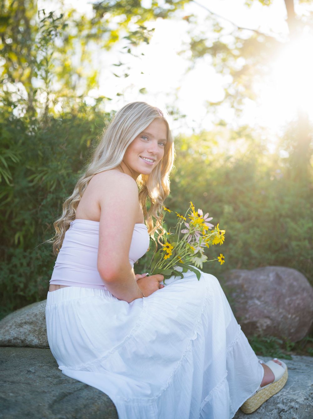Smiling blonde woman in white outfit holding wildflowers, sitting on rocks with sunlit greenery in background.