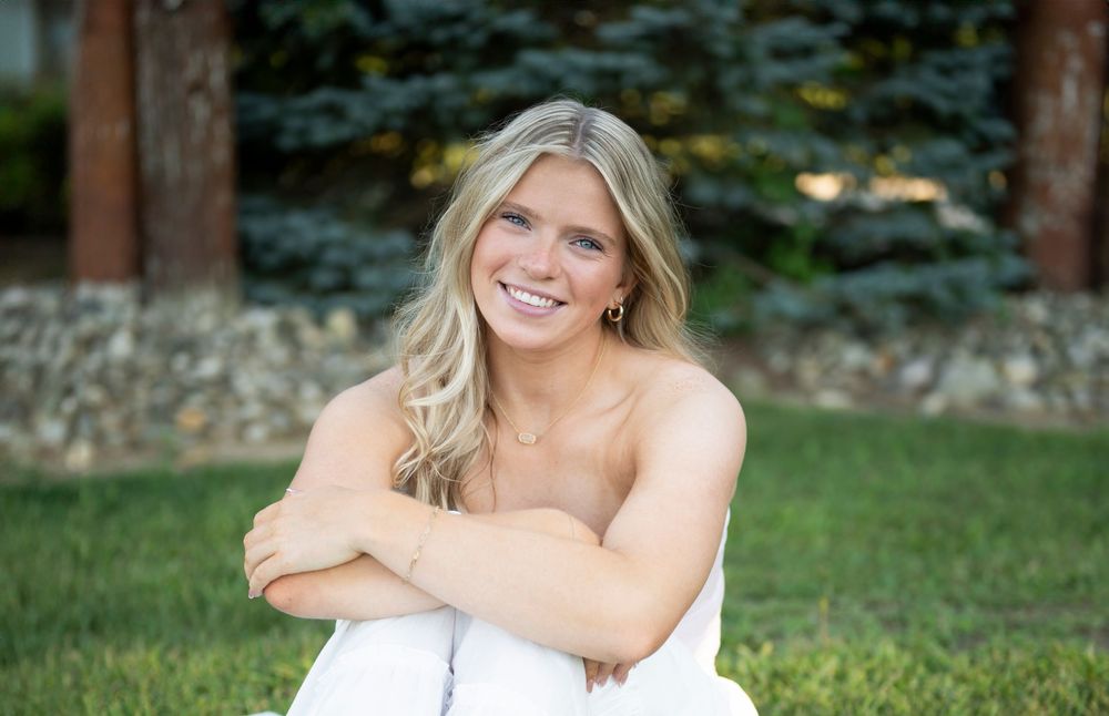 Smiling young blonde woman sitting on grass in white outfit with trees and stone wall background.