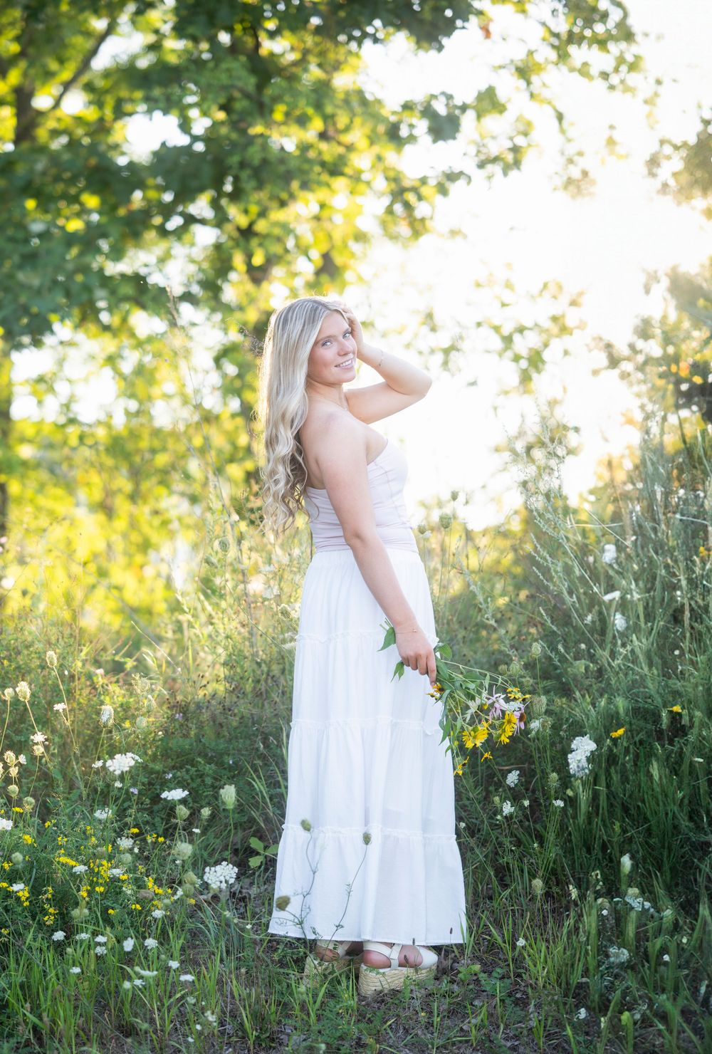 Blonde woman in white maxi dress holding wildflowers in a sunlit meadow surrounded by lush green trees.