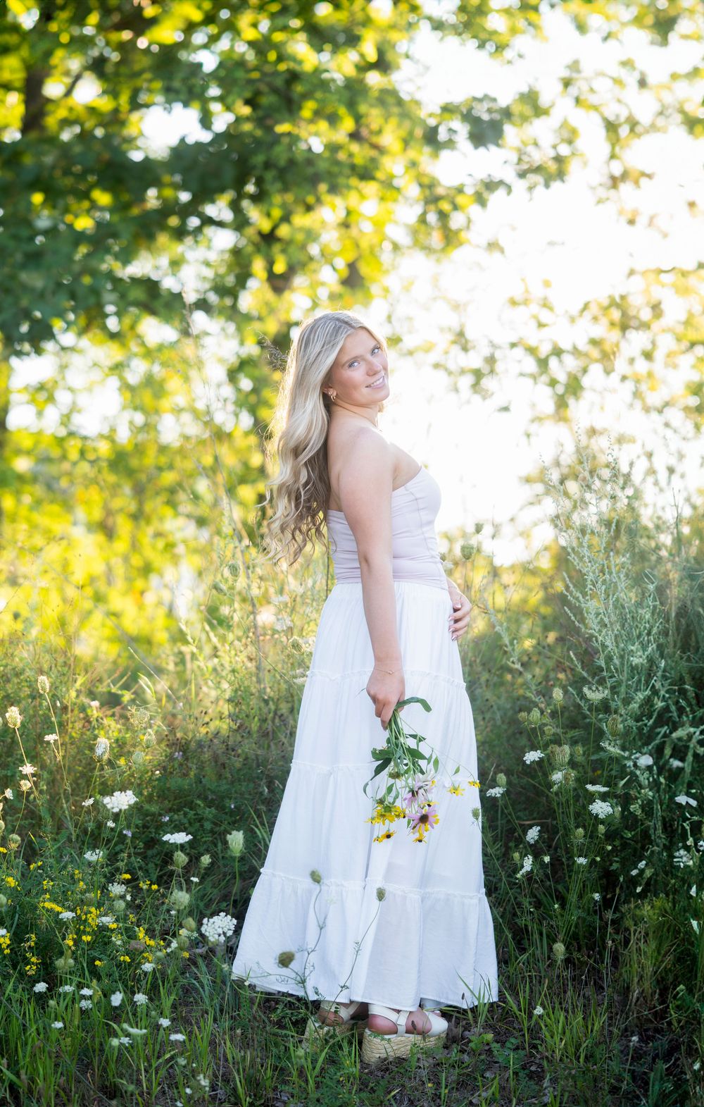 Blonde woman in white maxi dress holding wildflowers in a sunlit meadow.