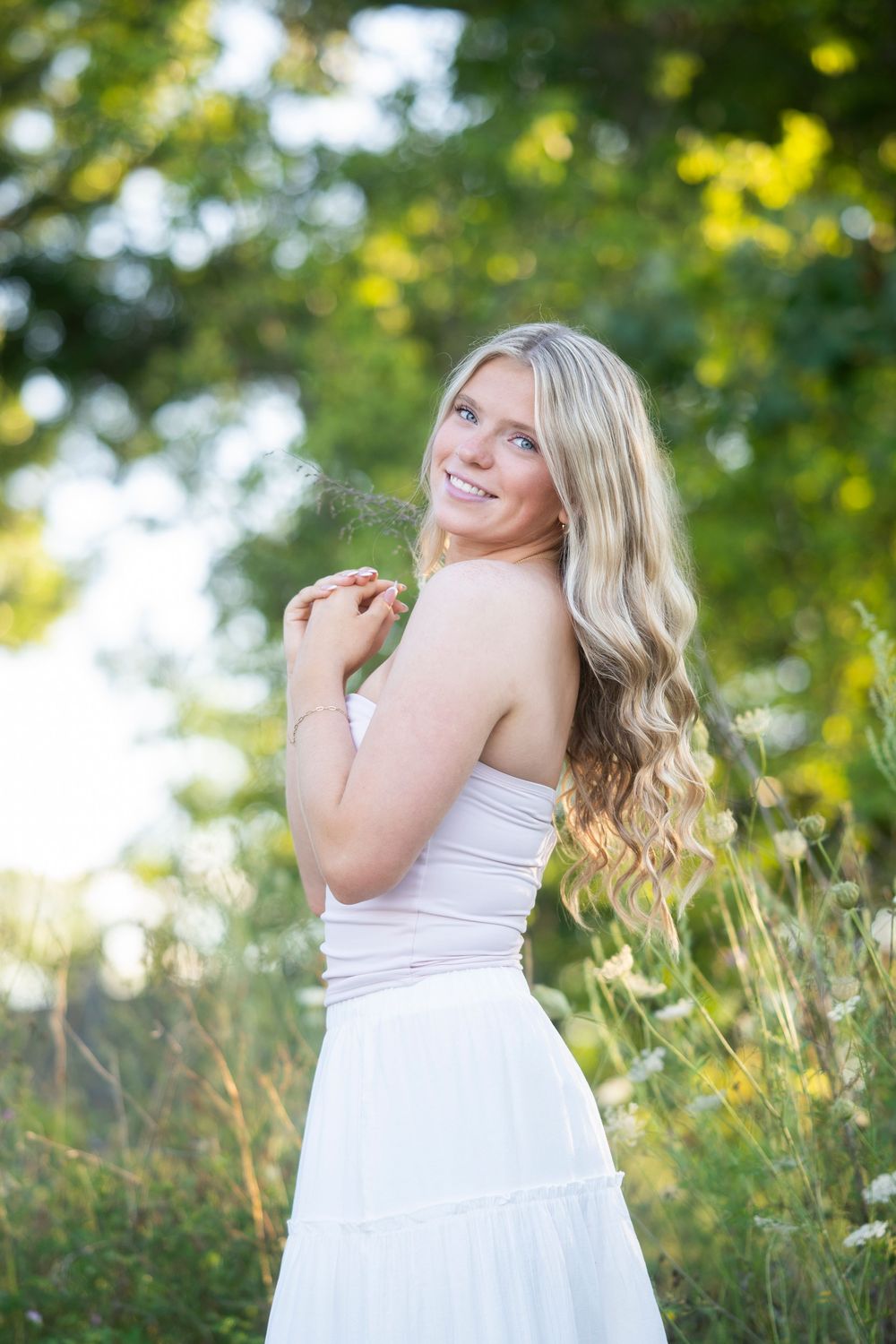 Smiling blonde woman in white dress standing in sunlit green field, looking over shoulder with long wavy hair.