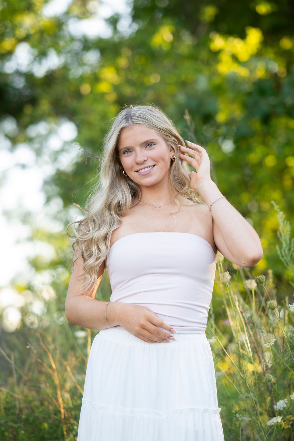 Smiling blonde woman in white strapless dress standing in a sunlit garden with green trees.