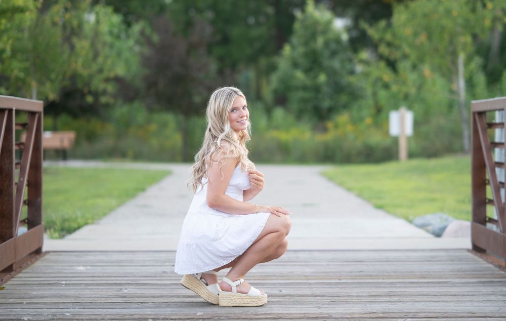 Blonde woman in white dress crouching on wooden bridge in a lush green park, smiling over her shoulder.