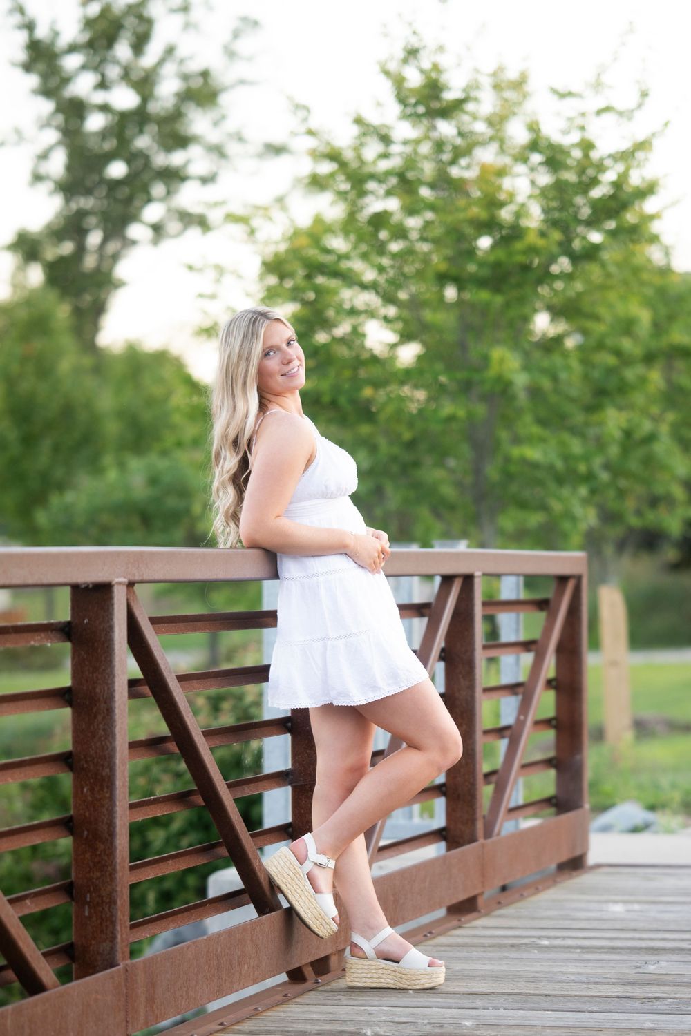 Young blonde woman in white sundress and platform sandals posing on a wooden bridge surrounded by lush green trees.