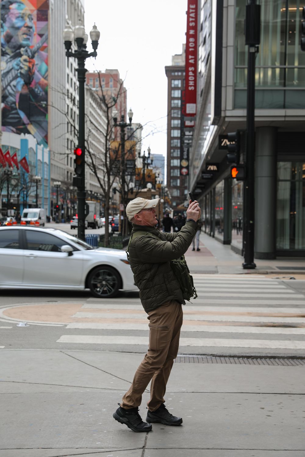 Man in green jacket takes a selfie on a busy urban street corner with city buildings and traffic in the background.