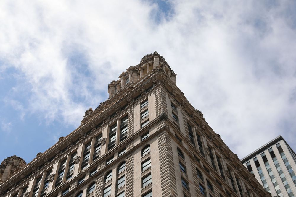 Historic ornate skyscraper corner viewed from below against a partly cloudy blue sky in an urban city setting.