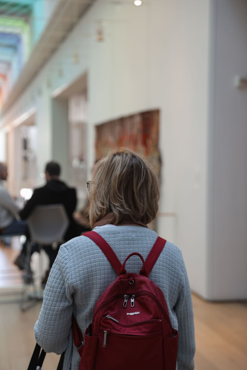 Woman with red backpack and gray jacket viewed from behind inside an art museum gallery.