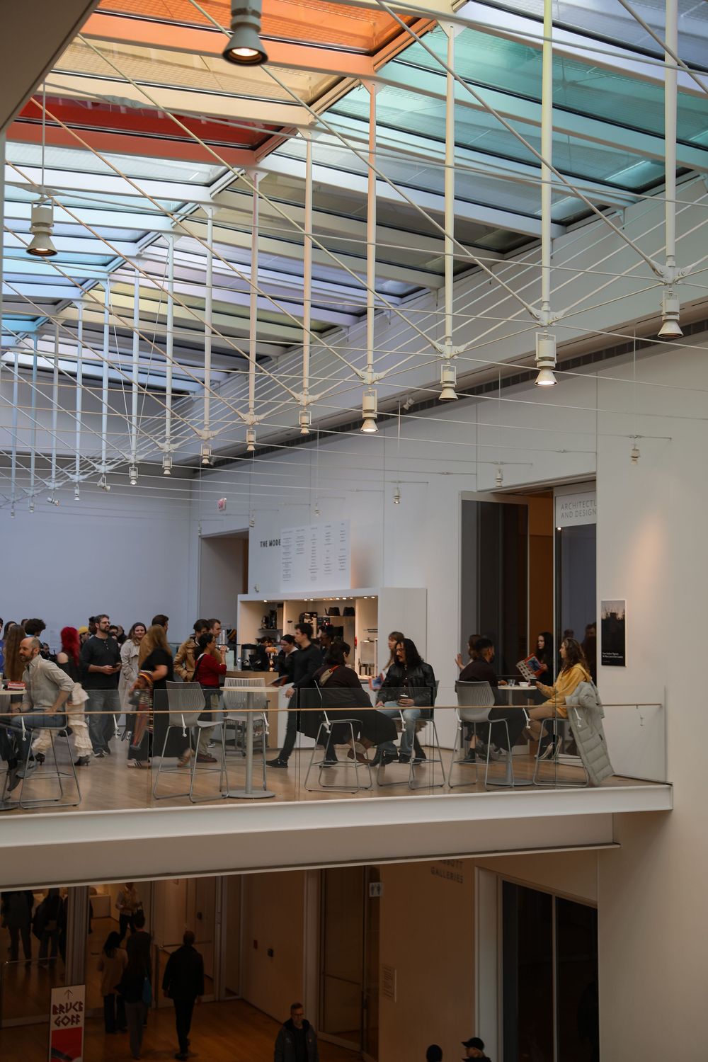 Modern museum atrium with colorful glass ceiling, visitors dining on upper balcony level, white walls and open space below.