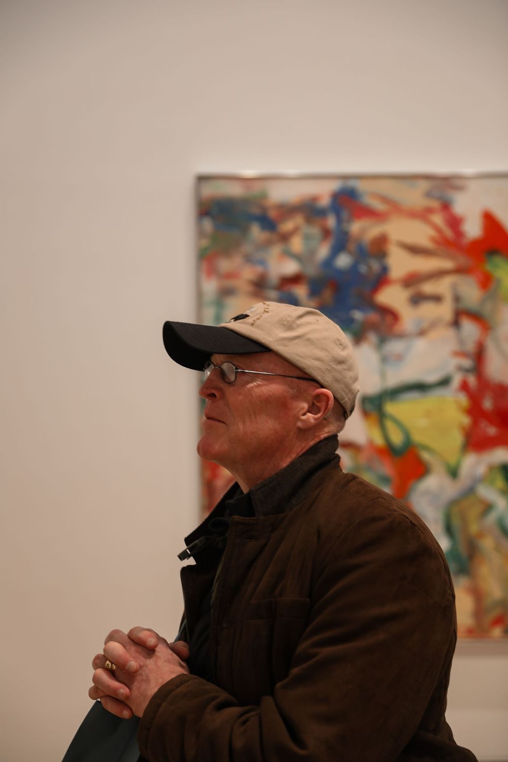 Elderly man in a baseball cap and glasses sits contemplatively in an art gallery with colorful abstract painting behind him.
