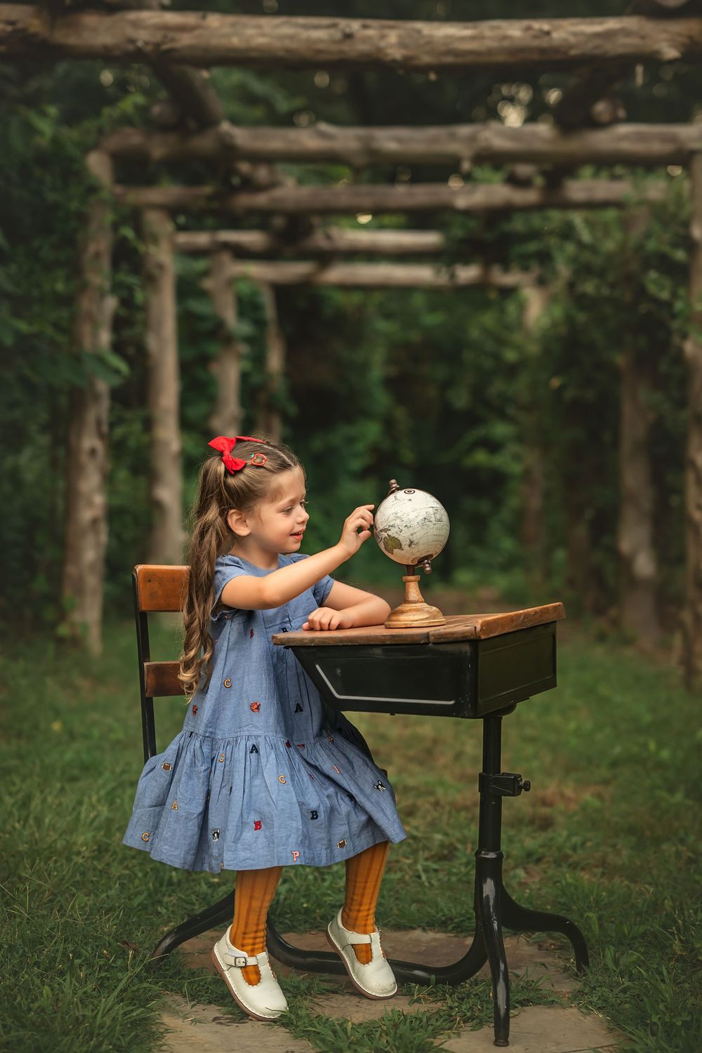 Young girl in blue dress exploring a vintage globe at an antique school desk outdoors under a wooden arbor.