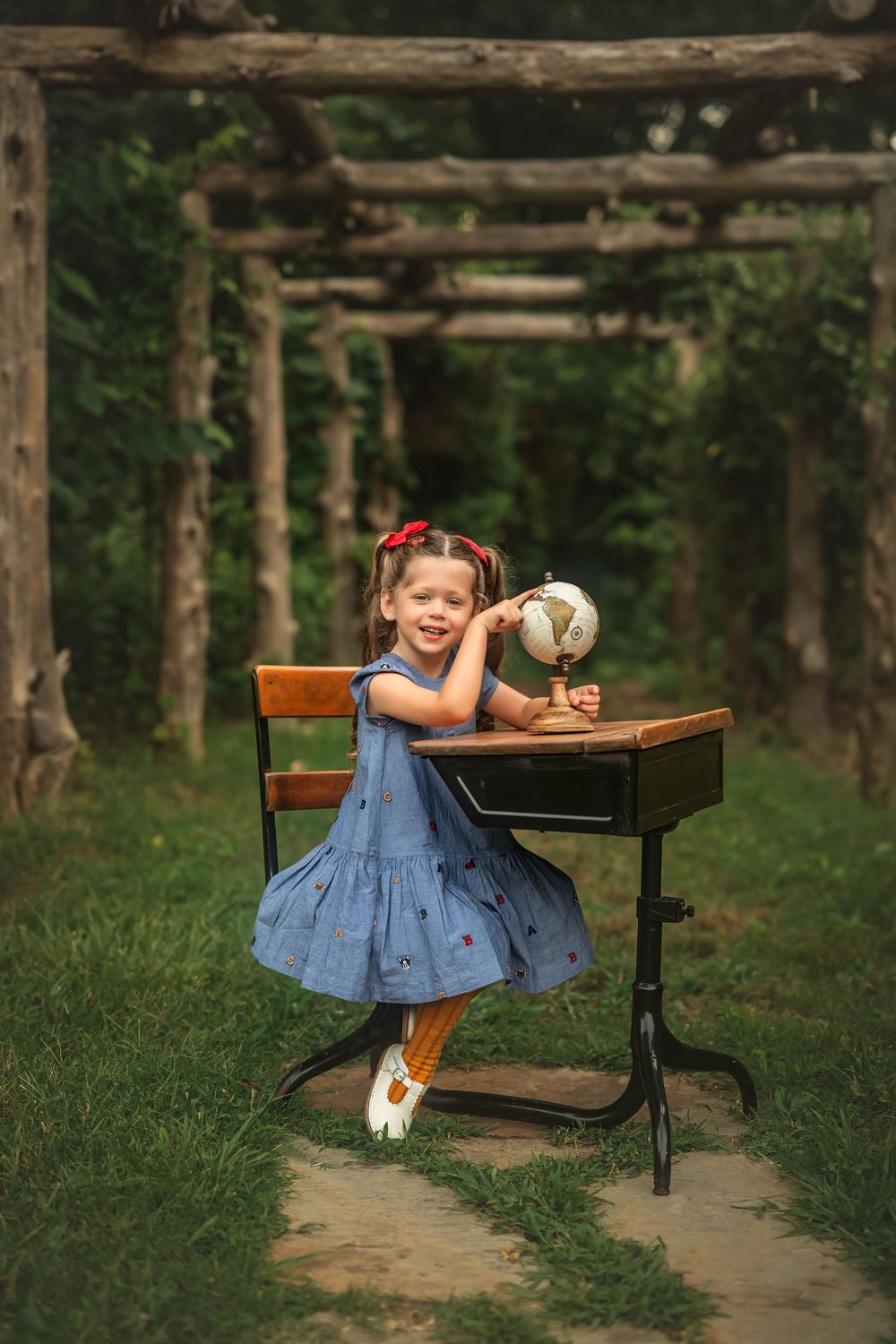Young girl in blue dress sits at vintage school desk outdoors, smiling with a globe, surrounded by green garden arbor.