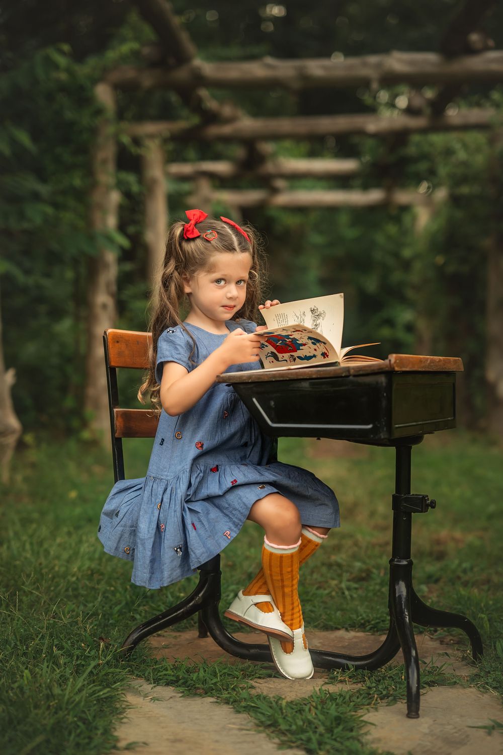 Young girl in blue dress and orange socks sits at vintage school desk outdoors, reading a book in a lush green garden.