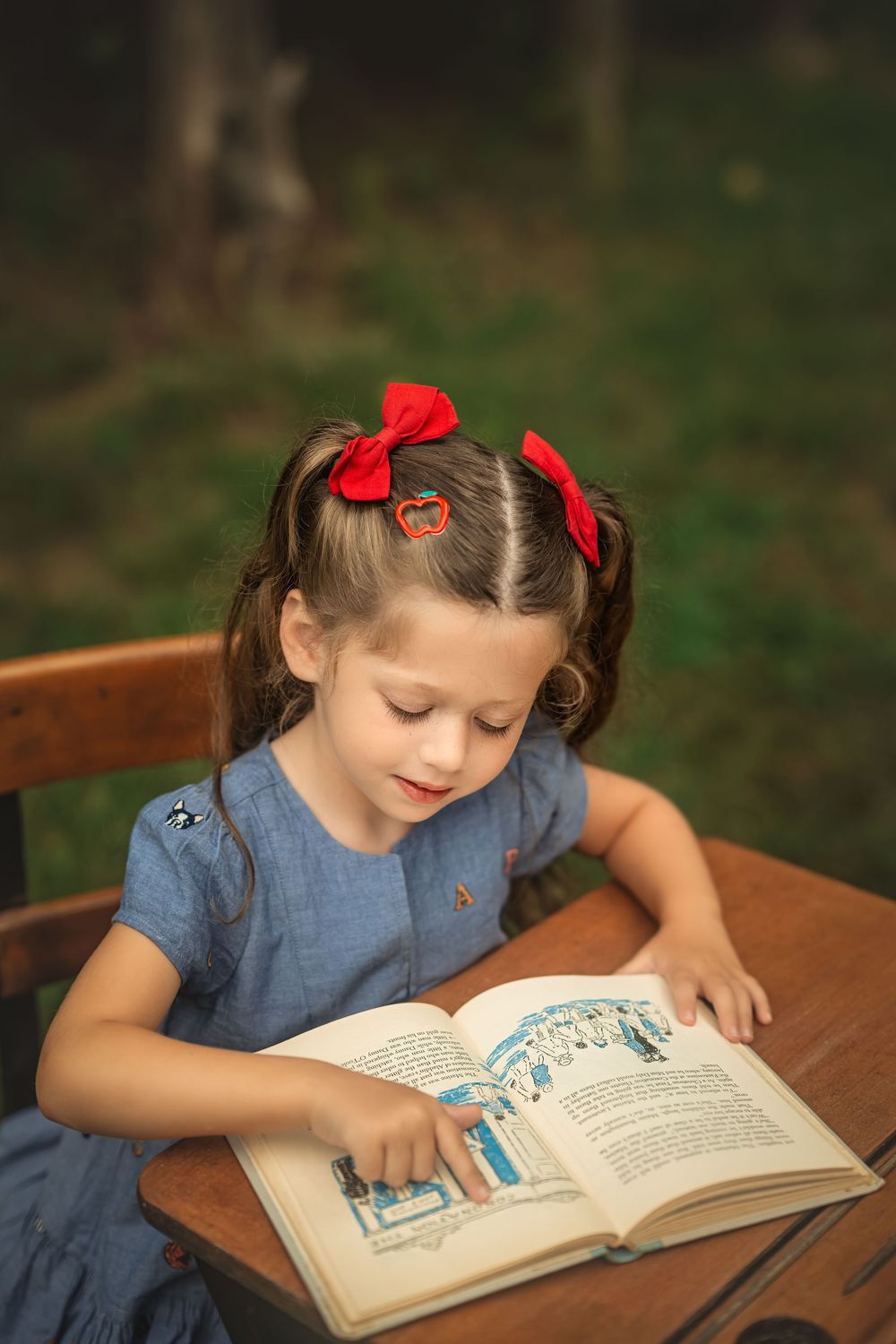 Young girl with red bows reading a book outdoors at a wooden desk, wearing a blue dress in a garden setting for back to school photos