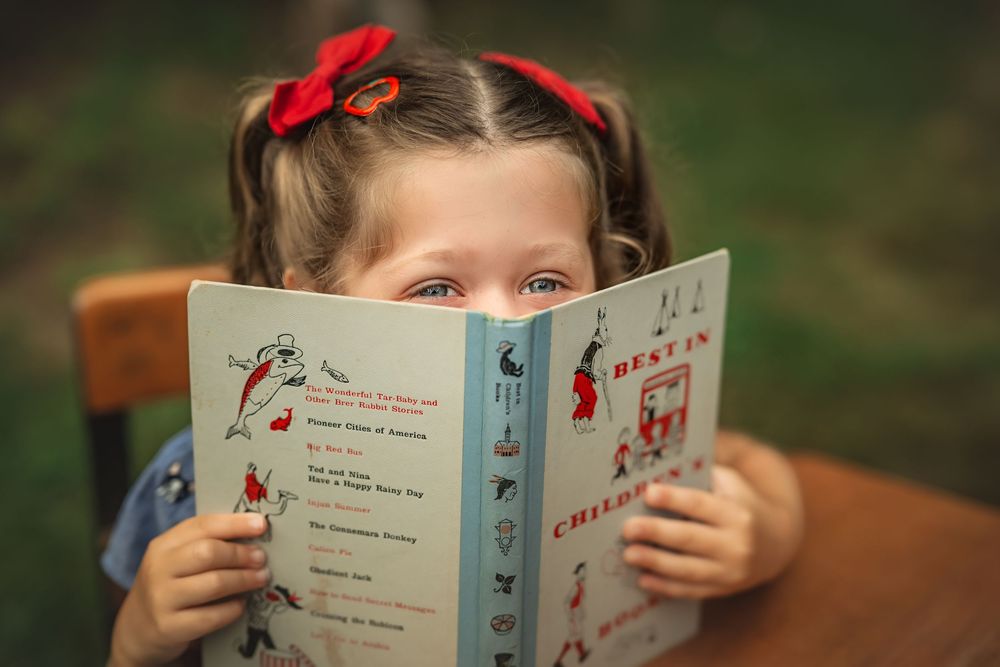 Young girl with red hair ribbons reads a colorful children's book outdoors, hiding her face behind the pages.