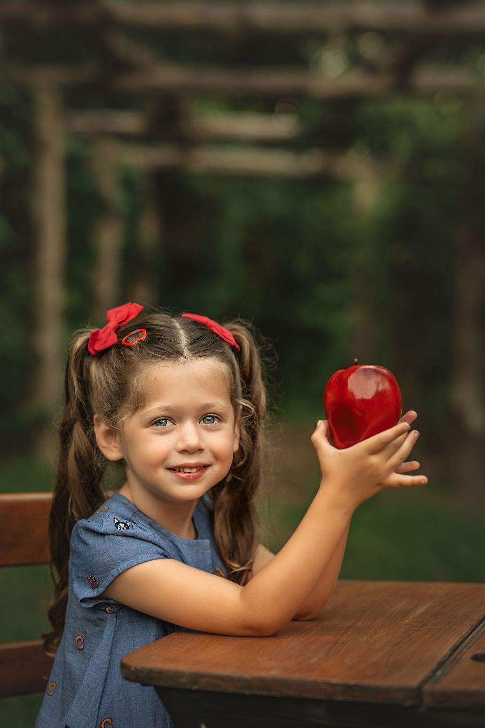Young girl smiling at a wooden table outdoors, holding up a shiny red apple, wearing a blue dress with red hair bows for back to school pics