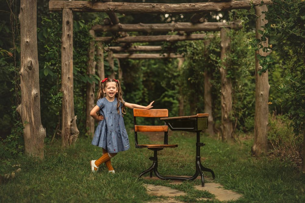 A young girl in a blue dress stands beside a vintage wooden school desk under a rustic log pergola in a lush garden.