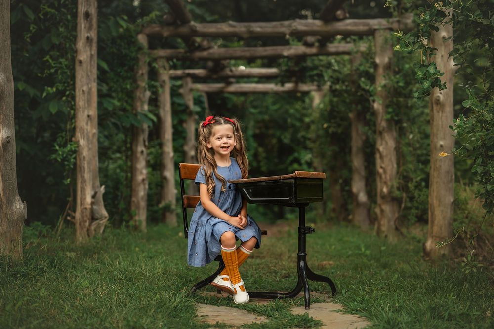 A smiling little girl in a blue dress and orange knee socks sits at an antique school desk outdoors in a lush green garden.
