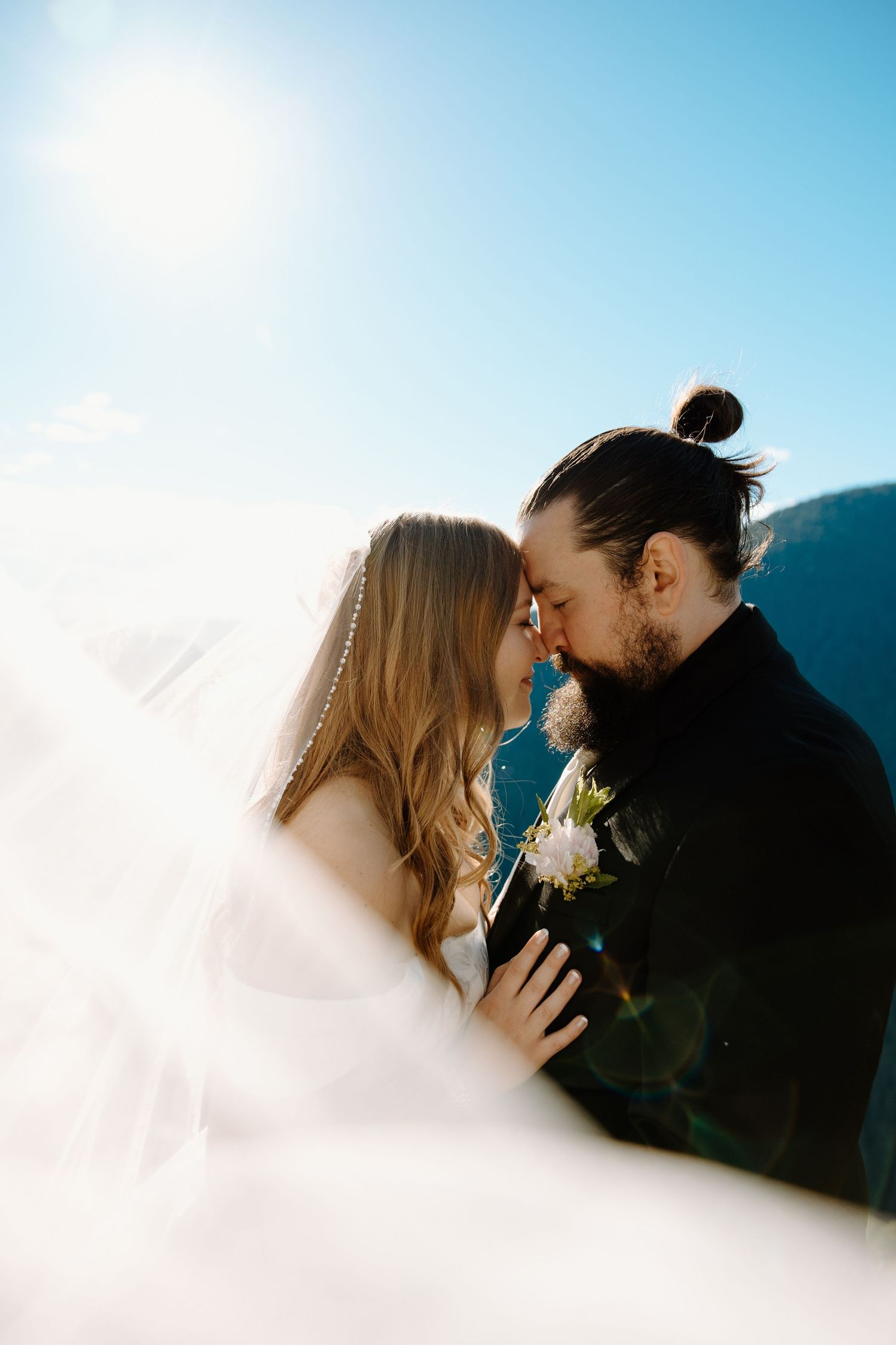 Bride and groom share a romantic kiss outdoors with flowing veil, mountain backdrop, and bright sunlight on their wedding day.