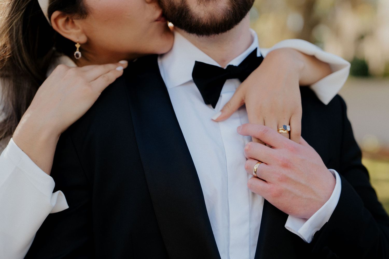 Bride and groom in elegant wedding attire, showcasing engagement ring and wedding bands during romantic embrace.
