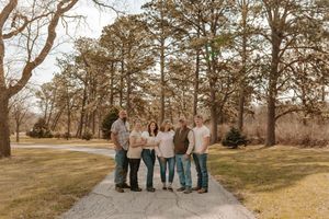 Family of six posing together on a gravel path surrounded by tall trees and green grass in a park setting.