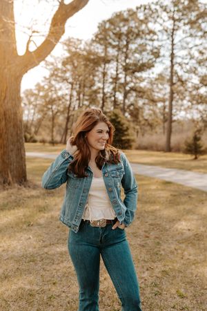 Young woman in denim jacket and jeans smiling outdoors at golden hour in a park setting.