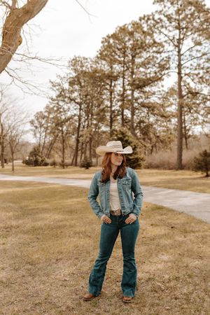 Woman in cowboy hat, denim jacket, and boots standing in a field with trees in the background.