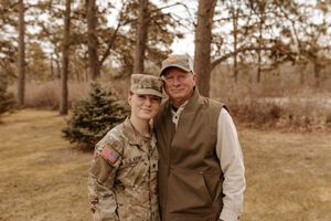 A soldier in military uniform poses with an older man in a vest outdoors near pine trees.