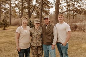 Family of four posing outdoors, including a woman in military camouflage uniform, smiling together near pine trees.