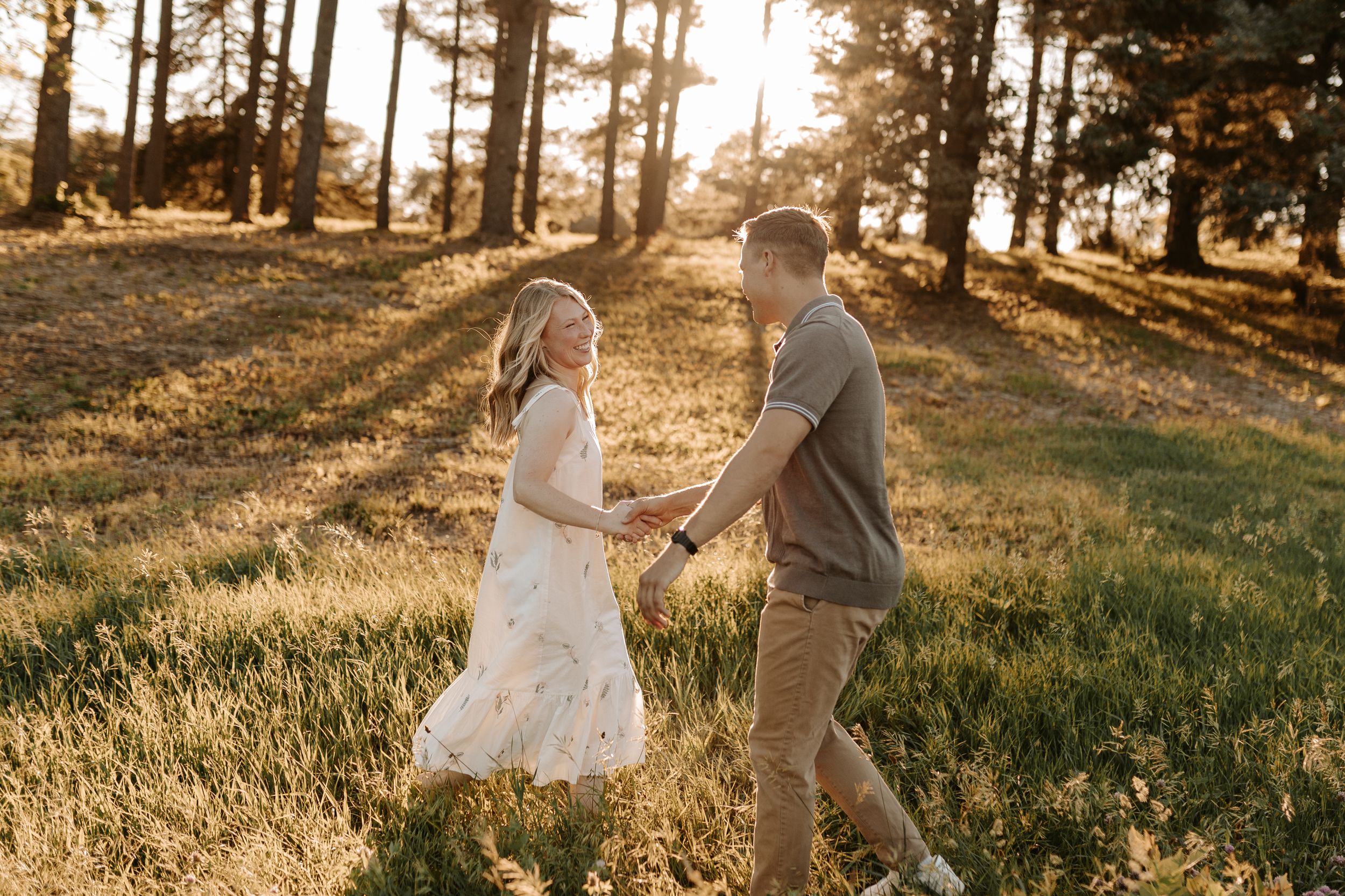 Couple holding hands dancing in golden sunlight among tall trees in a grassy field during a romantic outdoor engagement session.